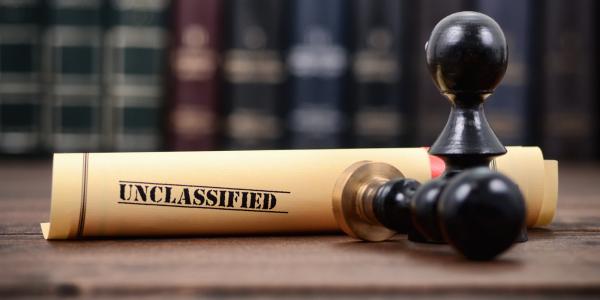 Image showing a rolled document labeled "UNCLASSIFIED" with a black rubber stamp on a wooden table, set against a backdrop of book spines.