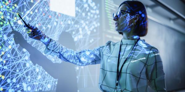 A presenter in a dark room pointing at a large screen displaying complex data visualizations.